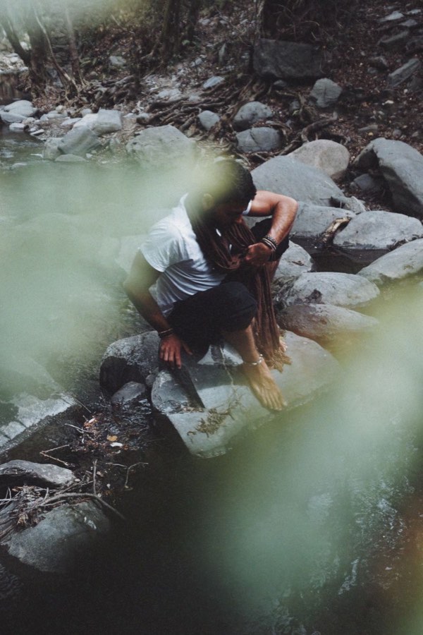 Stefano Bonanno agachado sobre una roca en un arroyo, con un pie en el agua, visto entre hojas desenfocadas.